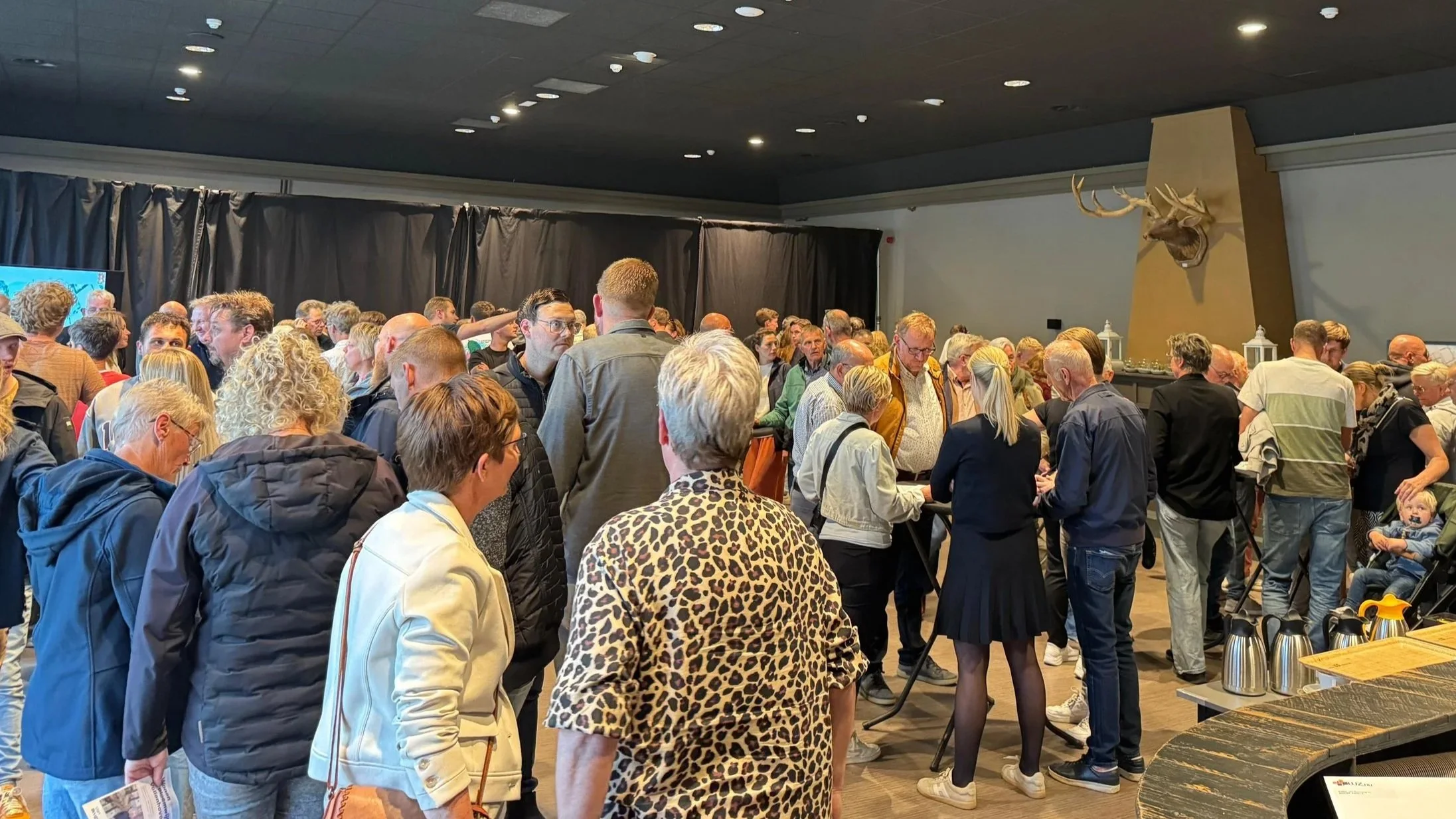 Een grote groep mensen wacht in rij in een kamervergadering of evenement, met een puntige muur en een hertenkop op de achtergrond.