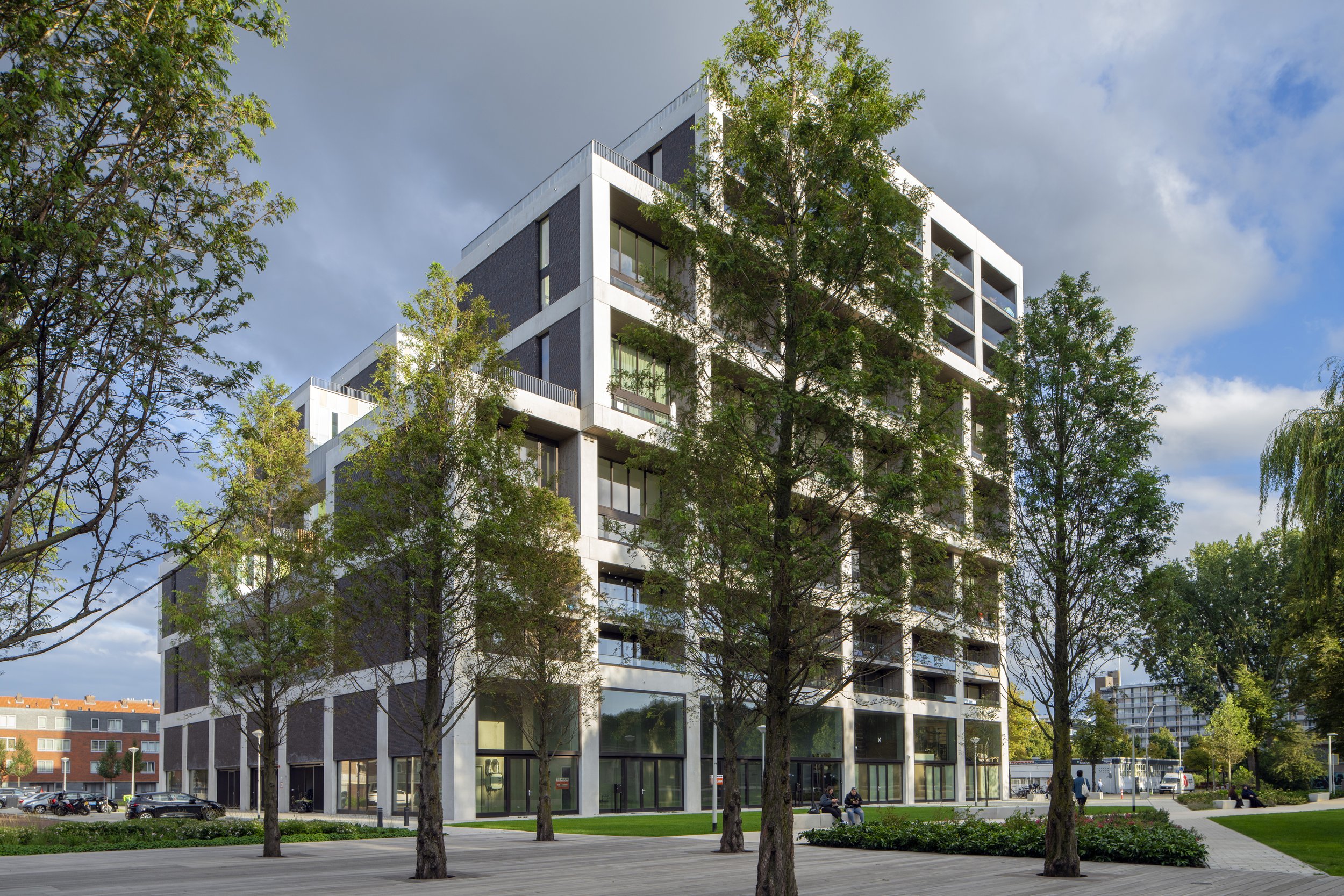 Modern, wit en zwarte kantoorgebouw met grote ramen, omringd door bomen en een park met banken en wandelpaden.