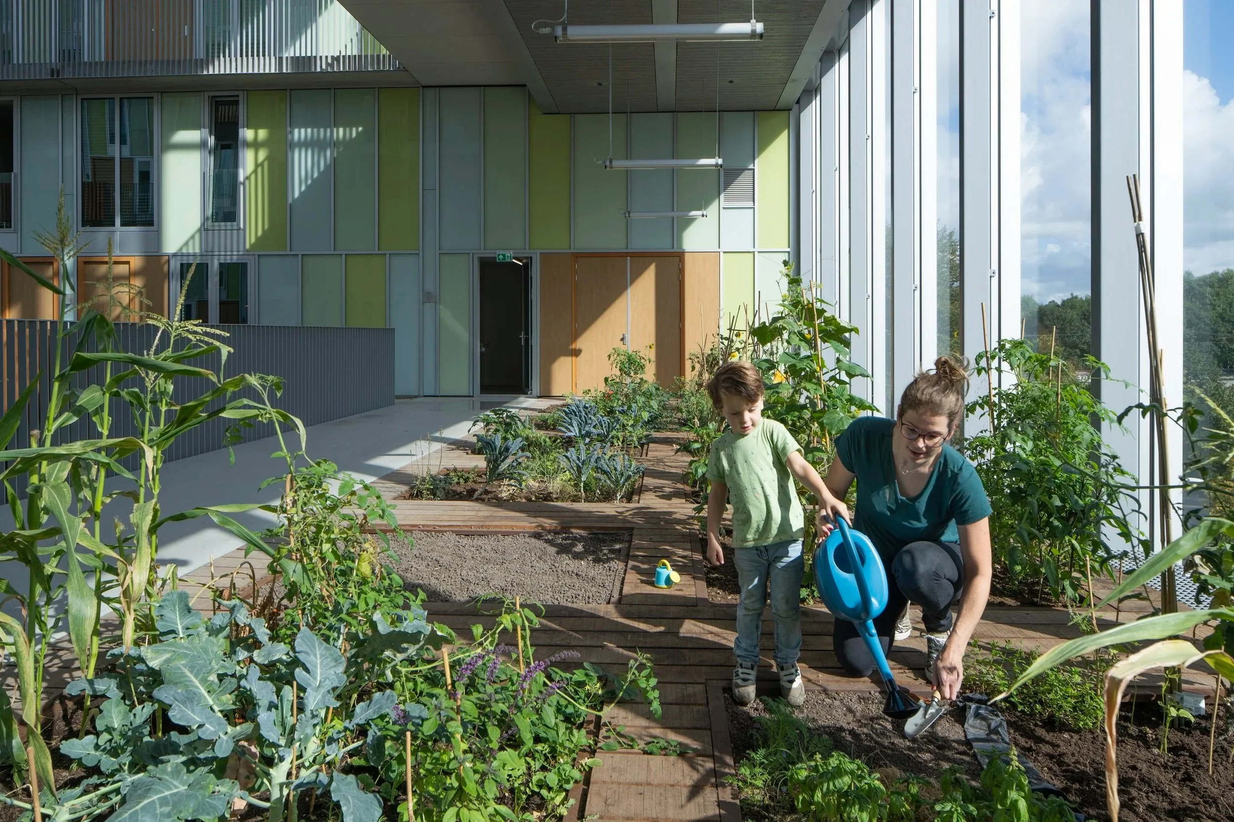 Een vrouw en een jongen die samen in een tuin binnen een gebouw tuinieren, met planten en een grote glazen muur op de achtergrond.