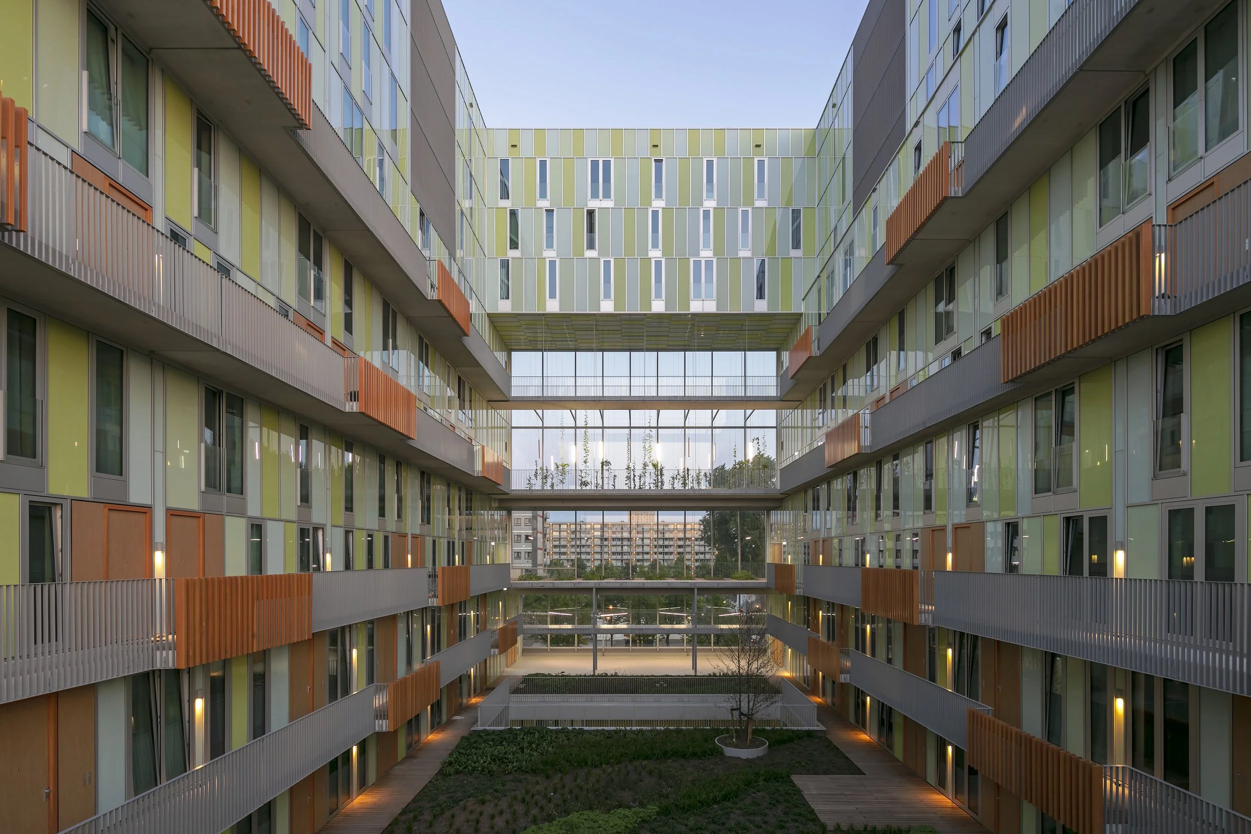 Binnenhof van een modern woningcomplex met meerdere verdiepingen, balkons met houten afwerkingen en een binnenplaats met groen en een boom, bij zonsondergang.