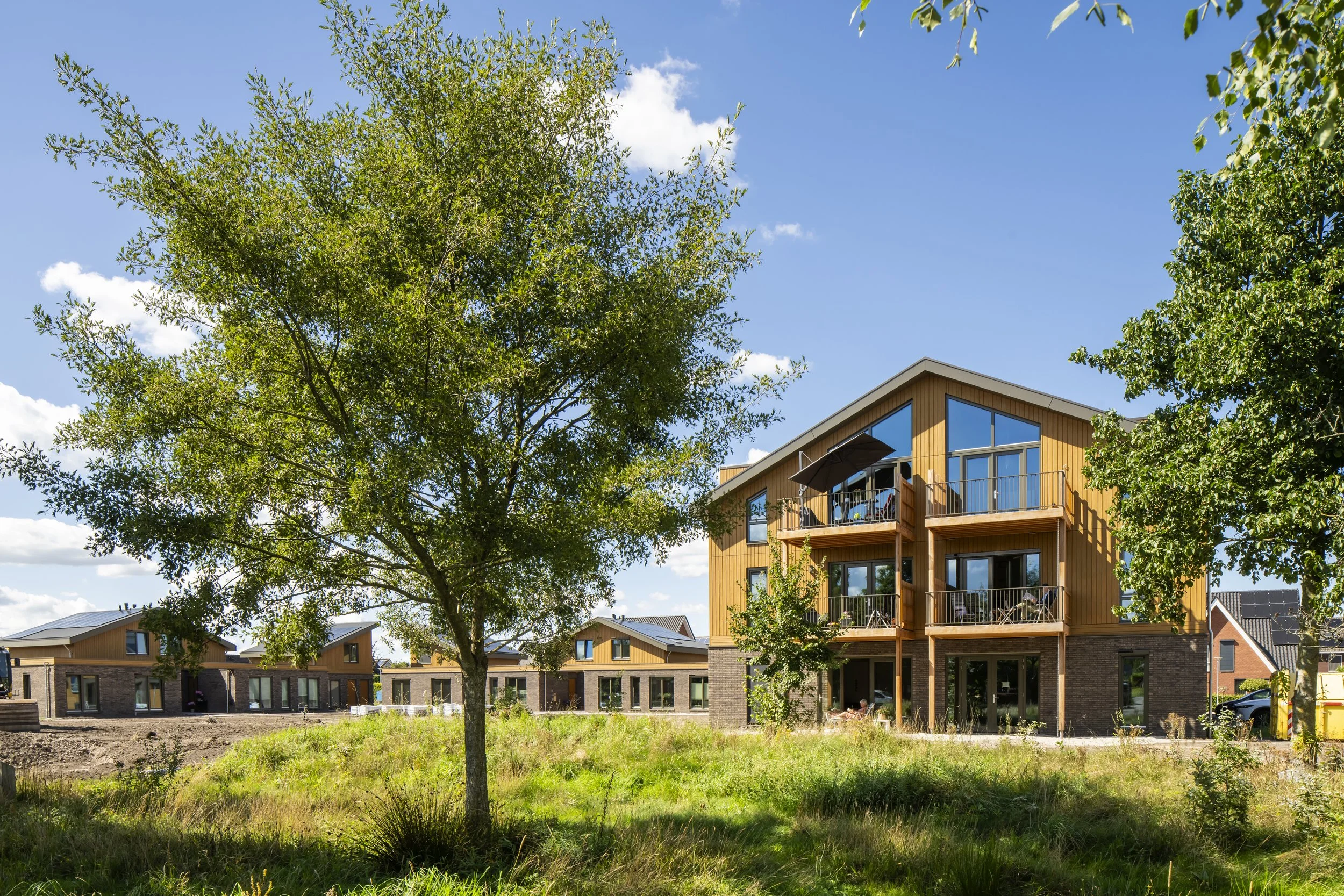 Moderne, houten drie verdiepingen tellend appartementengebouw met balkons, omgeven door groene bomen en een grasveld, onder een blauwe lucht met enkele witte wolken.