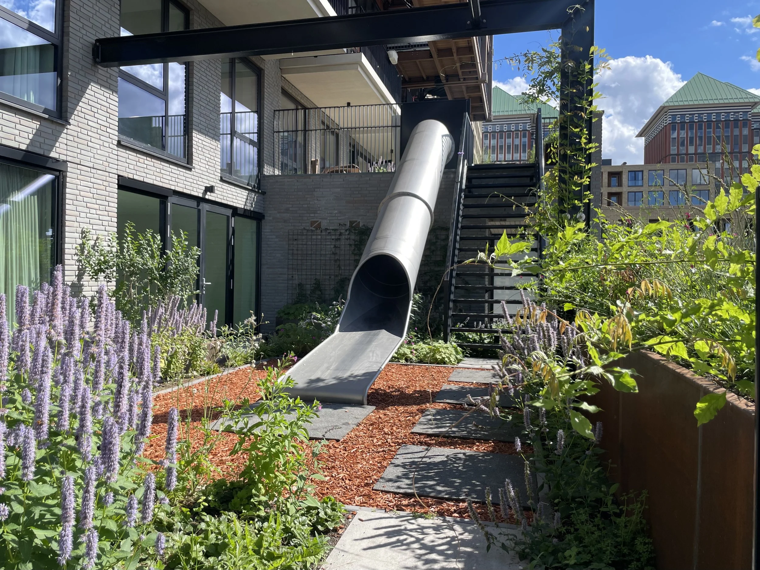 Bovenaanzicht van een moderne stadstuin met glijbaan, omgeven door groen, bloemen en planten, met flats in de achtergrond en een blauwe lucht met wolken.