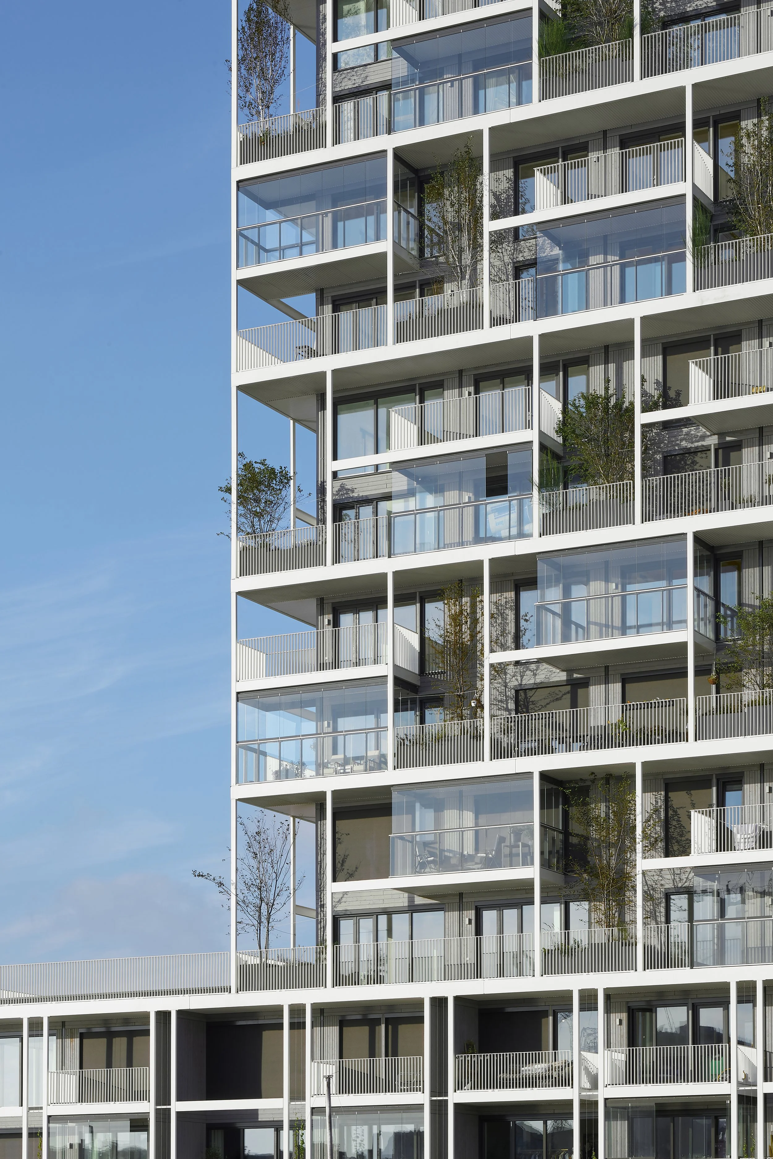 Moderne hoogbouw met glazen balustrades en balkons, enkele met planten, onder een heldere blauwe lucht.