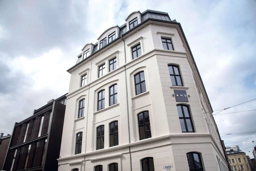 En hvit fløyelsbygning med seks etasjer og mørke vinduer, i en by med overskyet himmel.