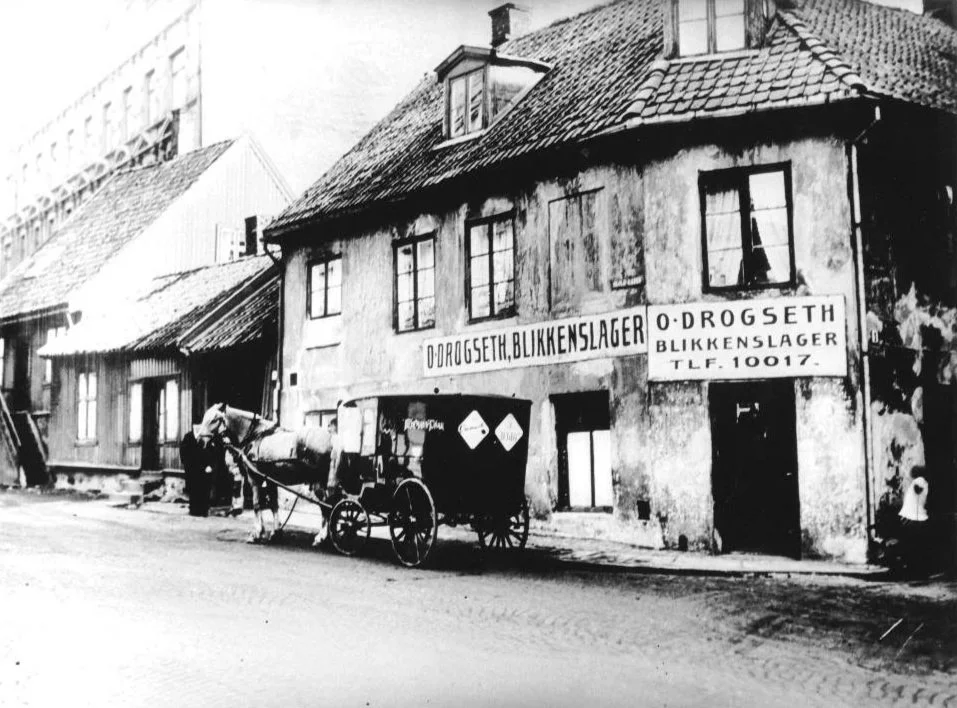 En svart-hvitt bilde av en gammel gate med et gammelt butikk- eller bedriftbygning med skilt som leser "Ø.DROGSETH, BLIKKENSLAGER". Det er en hest og vogn parkert foran bygningen, og en person står ved vognen, mens en annen står til venstre.