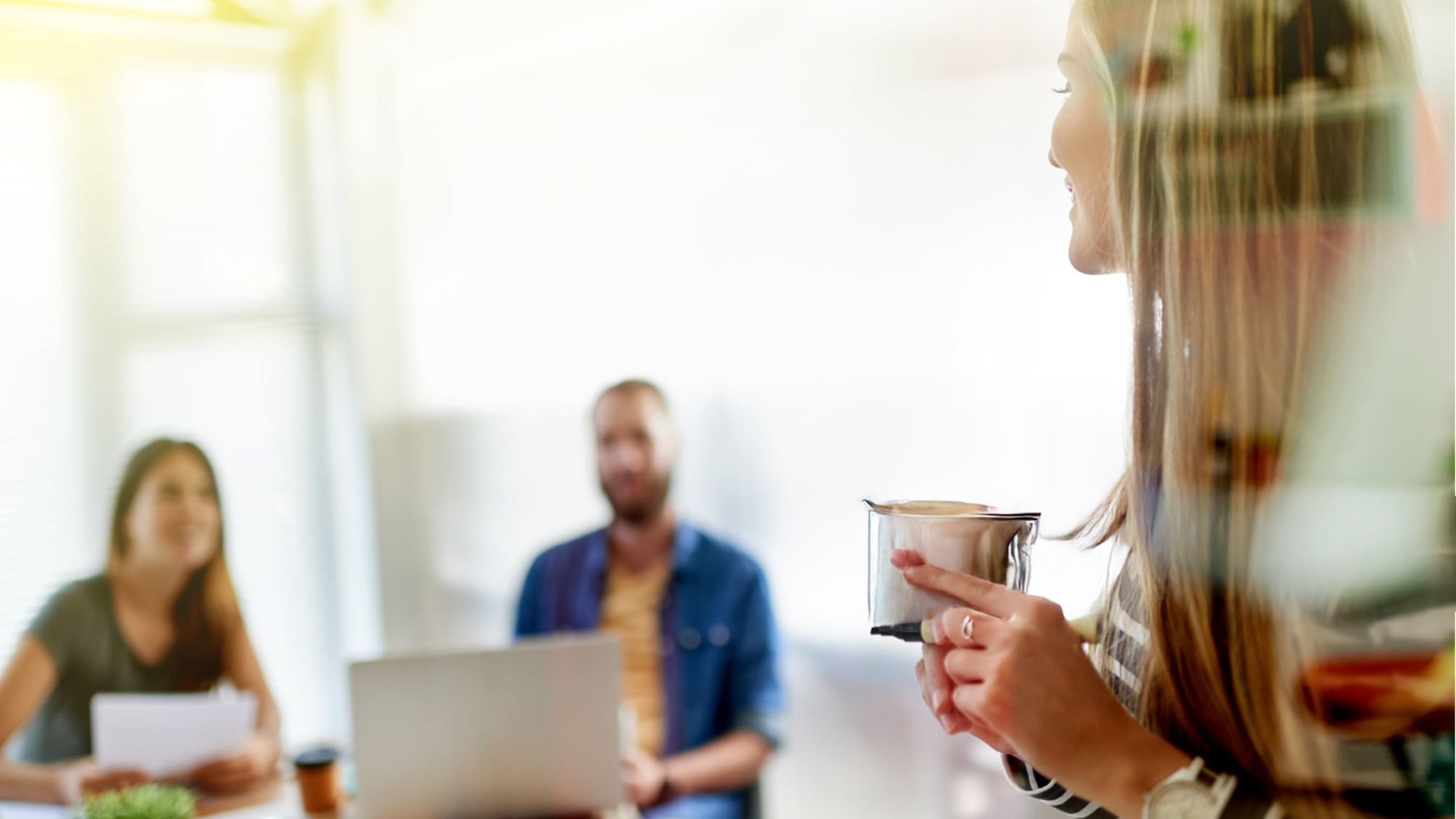 A woman with long blonde hair holding a ceramic mug, facing a group of two people in an office setting. Two colleagues, a woman and a man, sit at a table with a laptop and papers, smiling and engaging with her. The background has bright sunlight coming through windows, creating a cheerful atmosphere.