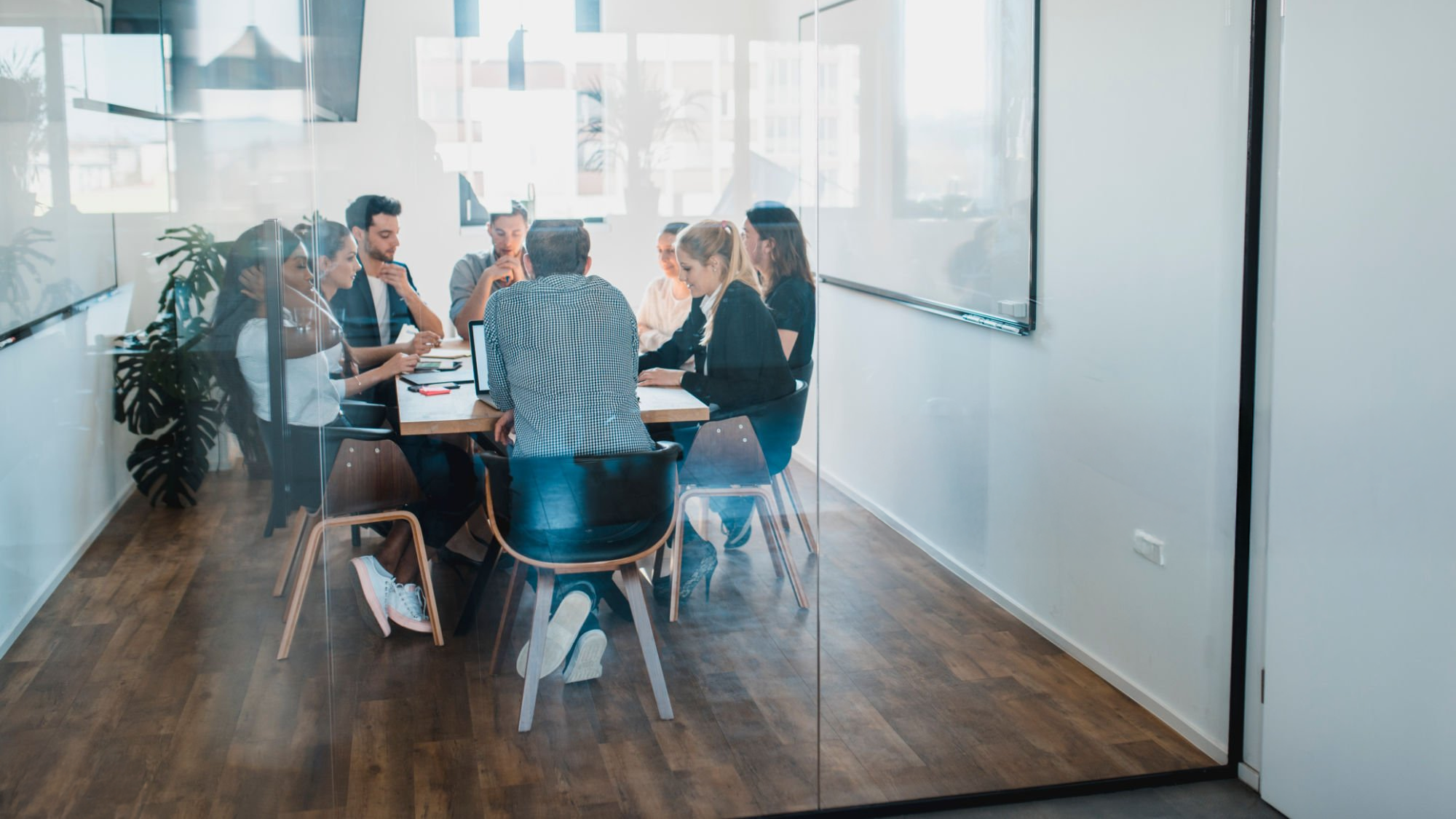 A group of colleagues in a business meeting seen through a glass wall in a modern office conference room with wooden floors, white walls, and large windows.