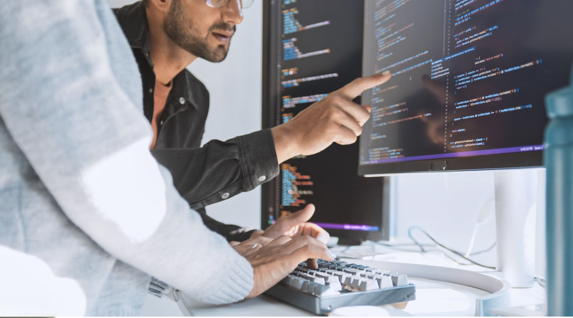 Two people working on a computer with a dual monitor setup, displaying lines of colorful code.