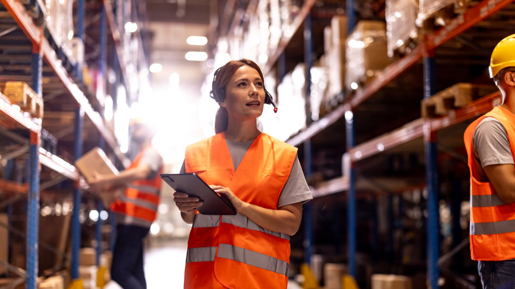 A woman wearing safety gear, including an orange reflective vest and a headset, holds a tablet while working in a warehouse with shelves loaded with boxes and goods.