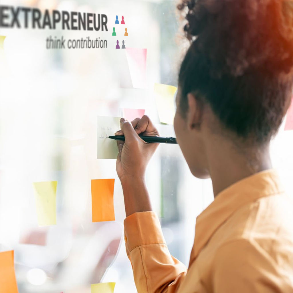 A person with curly hair and an orange shirt writing on post-it notes stuck to a window with the text 'EXTRAPRENEUR' and 'think contribution' on it.