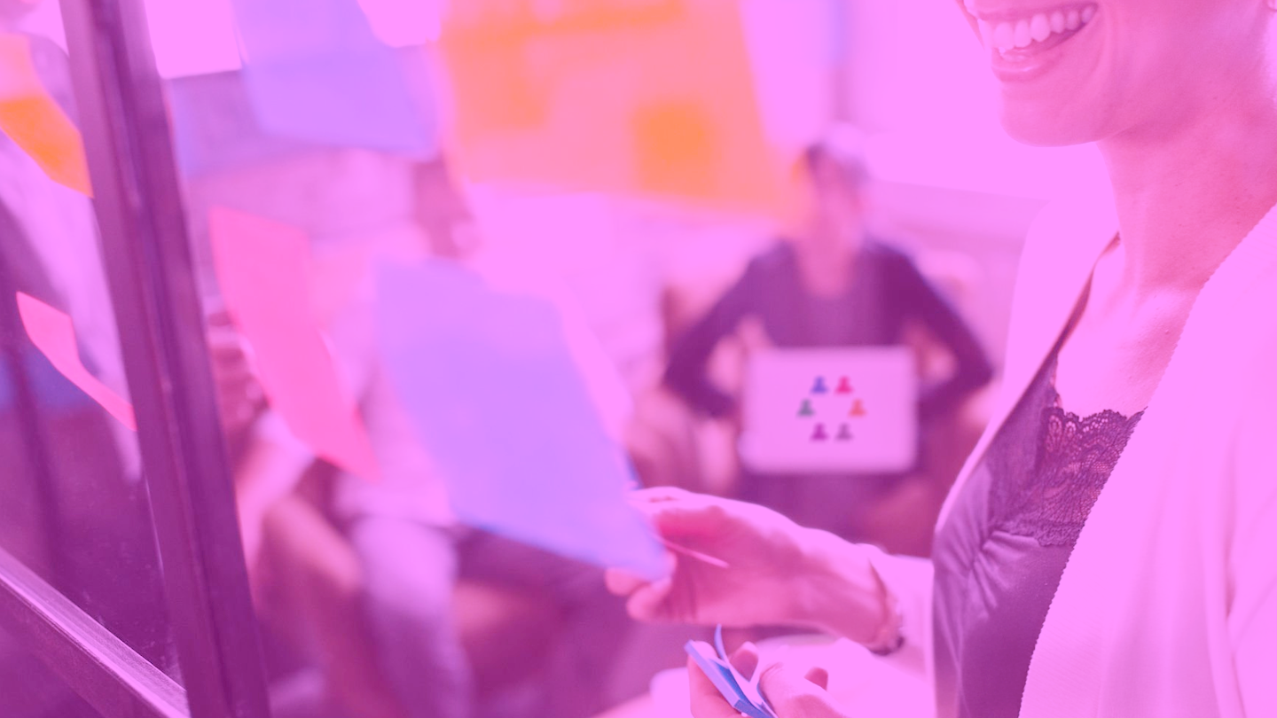 Close-up of a smiling woman wearing a black lace top and white blazer, paraphrased for accessibility, in a discussion with colleagues during a meeting.