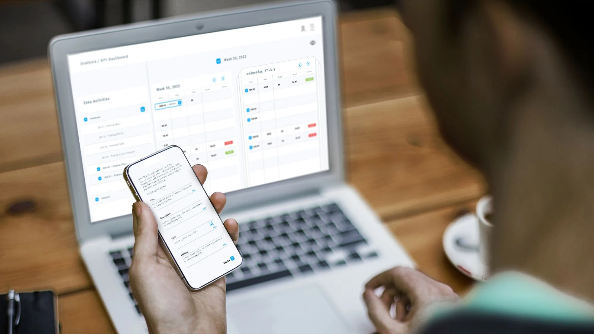Person holding smartphone with scheduling app open, looking at a laptop displaying a digital calendar on a wooden desk.
