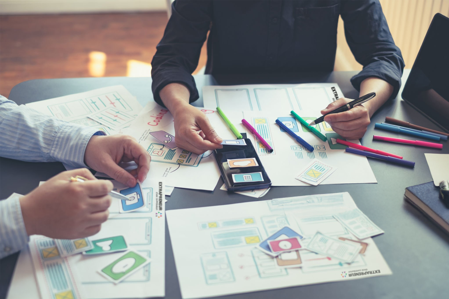 Two people working on a mobile app design with sketches, colored markers, and paper prototypes on a desk.