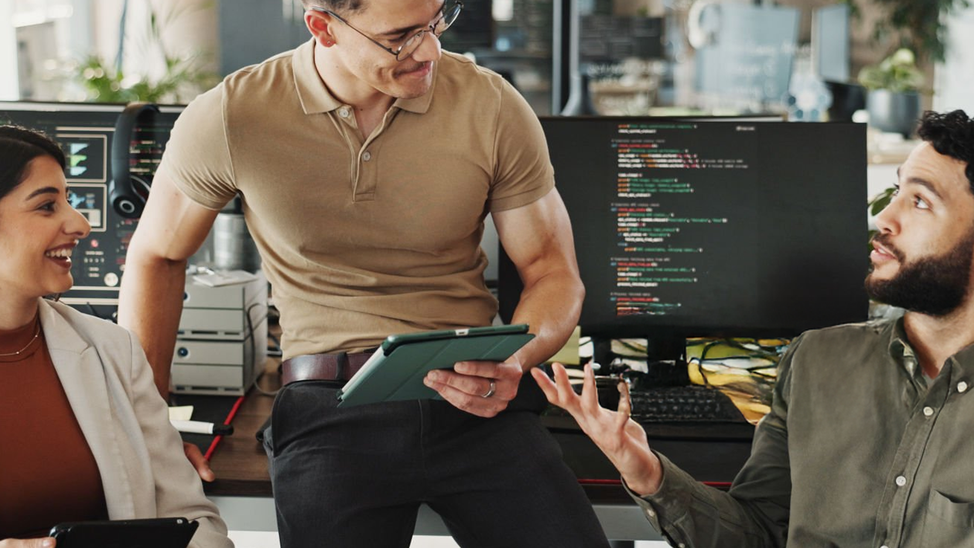 Three people in an office discussing, one holding a tablet, others smiling, computer screens with code in the background.