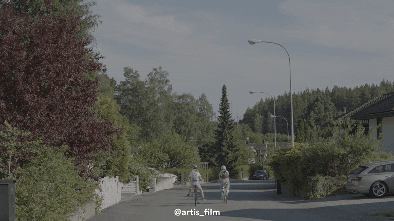 Two children riding bicycles on a suburban street with trees and parked cars on the sides.