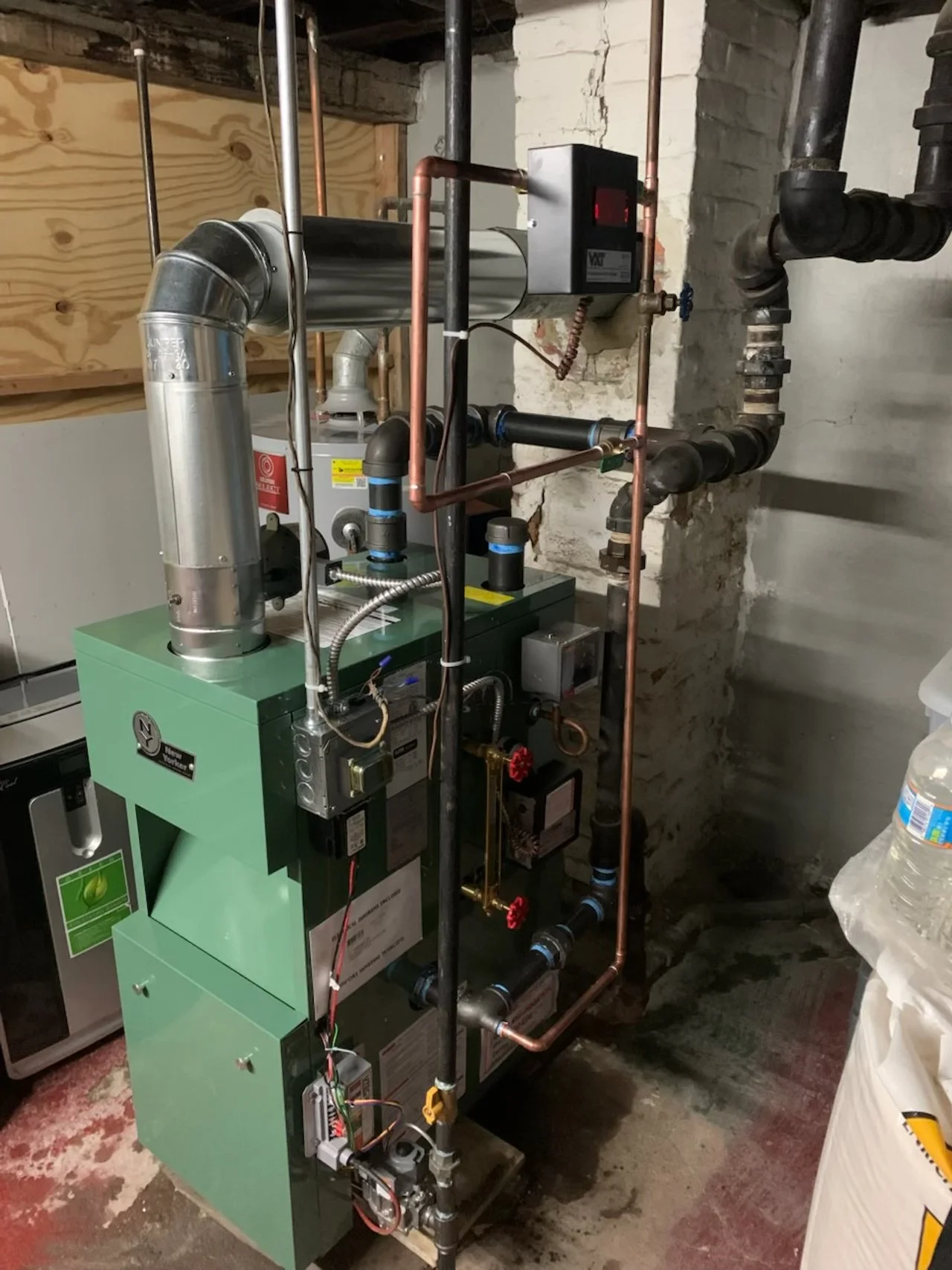 A green boiler or furnace with pipes and vents in a basement or utility room, surrounded by exposed brick and concrete walls.