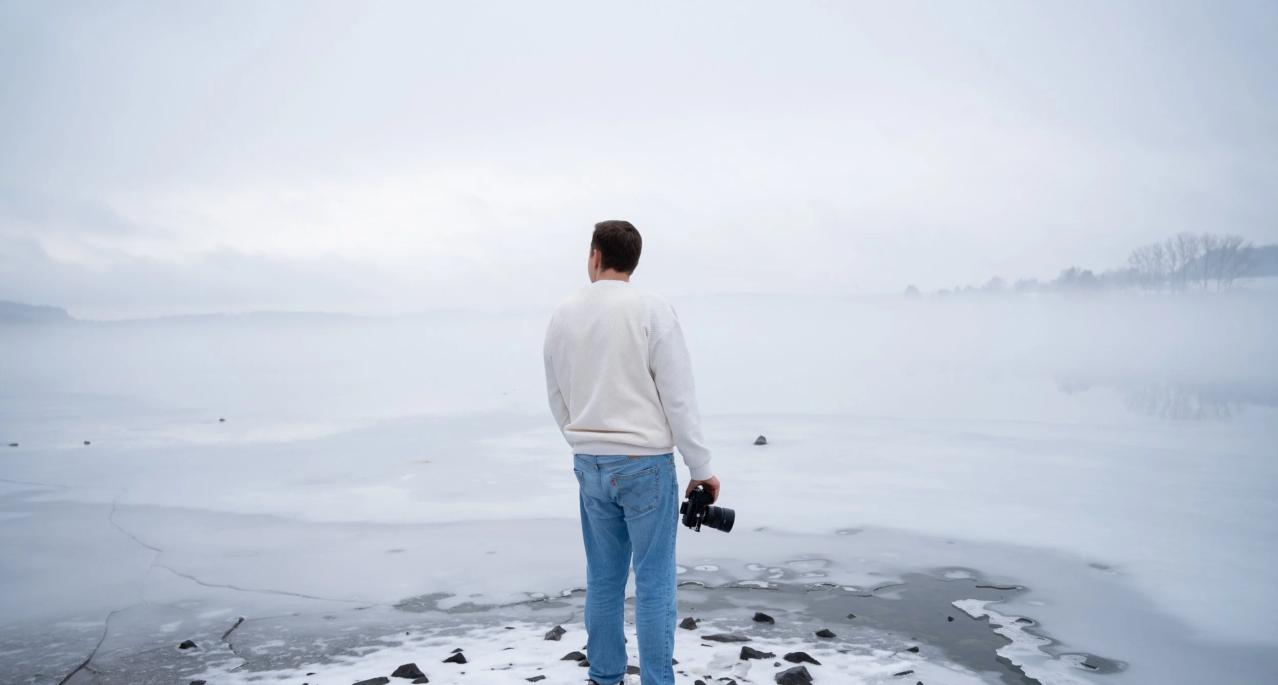 Mann steht am mit Eis bedeckten See, hält eine Kamera, Blick in die Ferne, nebliger Himmel, winterliche Szene.