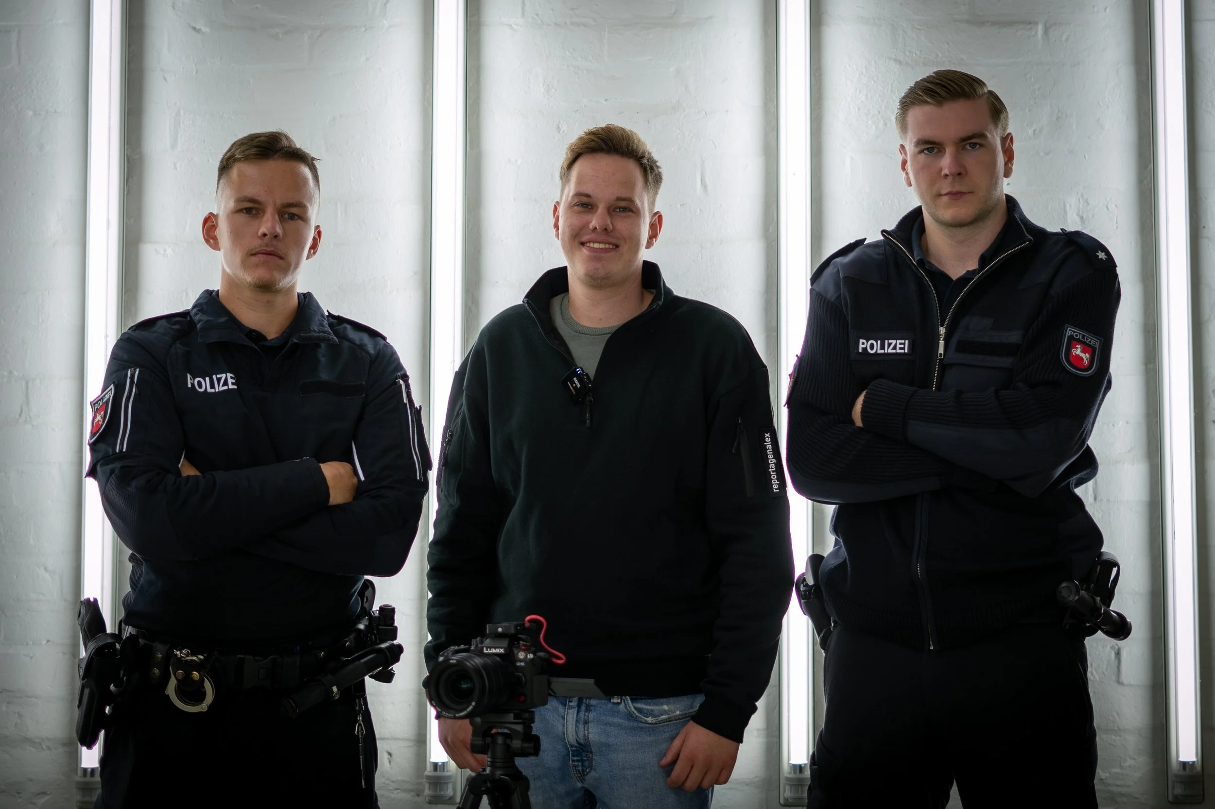 Zwei Polizisten in Uniform mit ausgestreckten Armen und ein Mann mit schwarzem Pullover, der vor einem professionellen Kamerastativ steht, im Hintergrund eine weiße Wand mit vertikalen Lichtleisten.