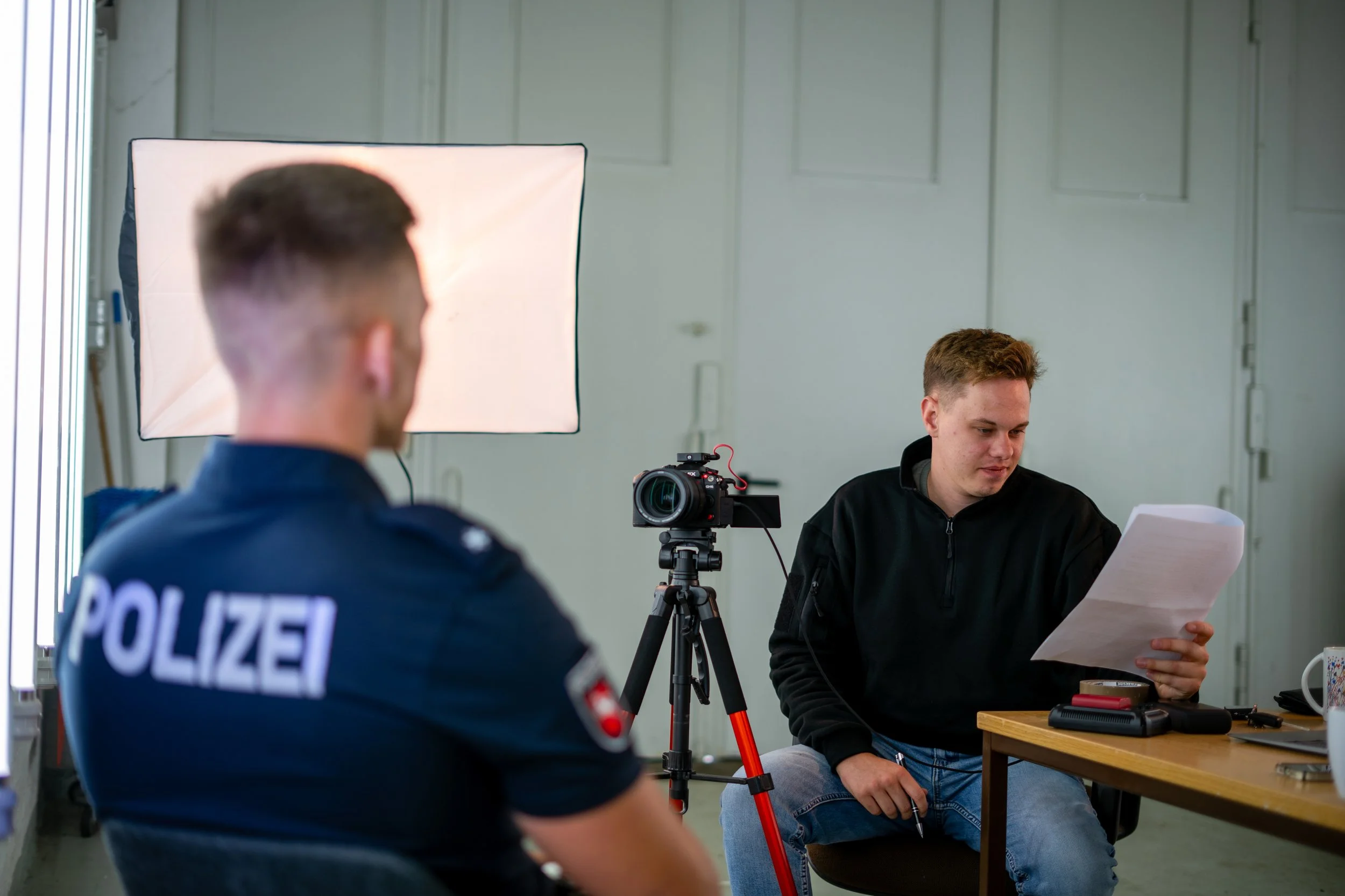 Zwei Männer, einer trägt eine Polizei-Uniform, sitzen in einem Raum, während ein junger Mann auf der anderen Seite einen Bericht liest, mit einer Kamera auf einem Stativ davor.