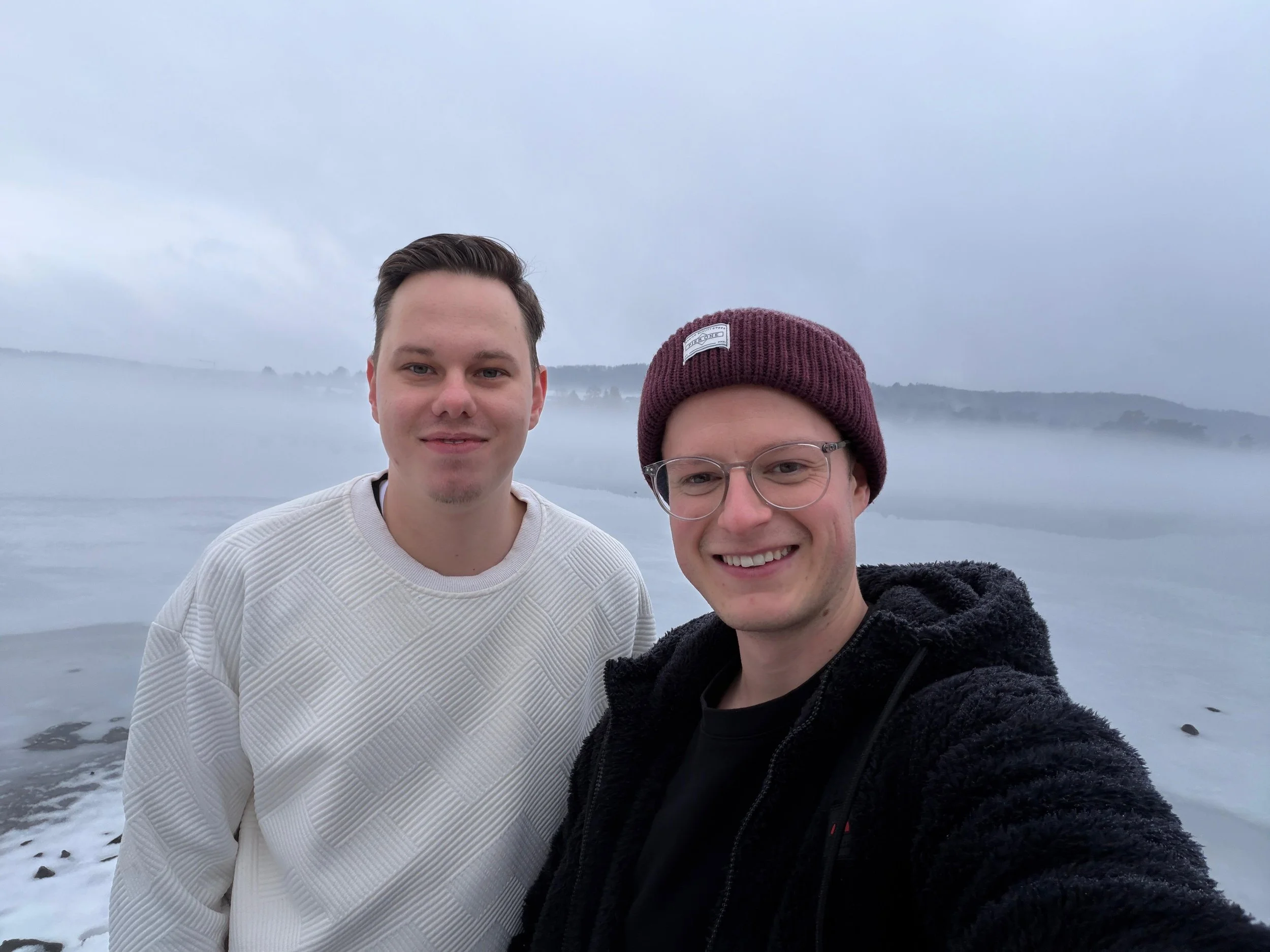 Zwei junge Männer machen ein Selfie am Fluss mit Nebel und bewölktem Himmel im Hintergrund.