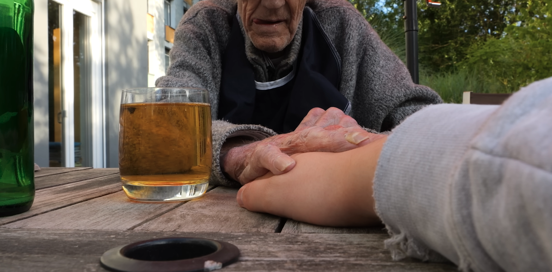 Ein älterer Mann hält die Hand einer jüngeren Person auf einem Holzgarten-Tisch, im Vordergrund steht ein Glas mit goldenem Getränk. Im Hintergrund sind Bäume und Garten zu sehen.