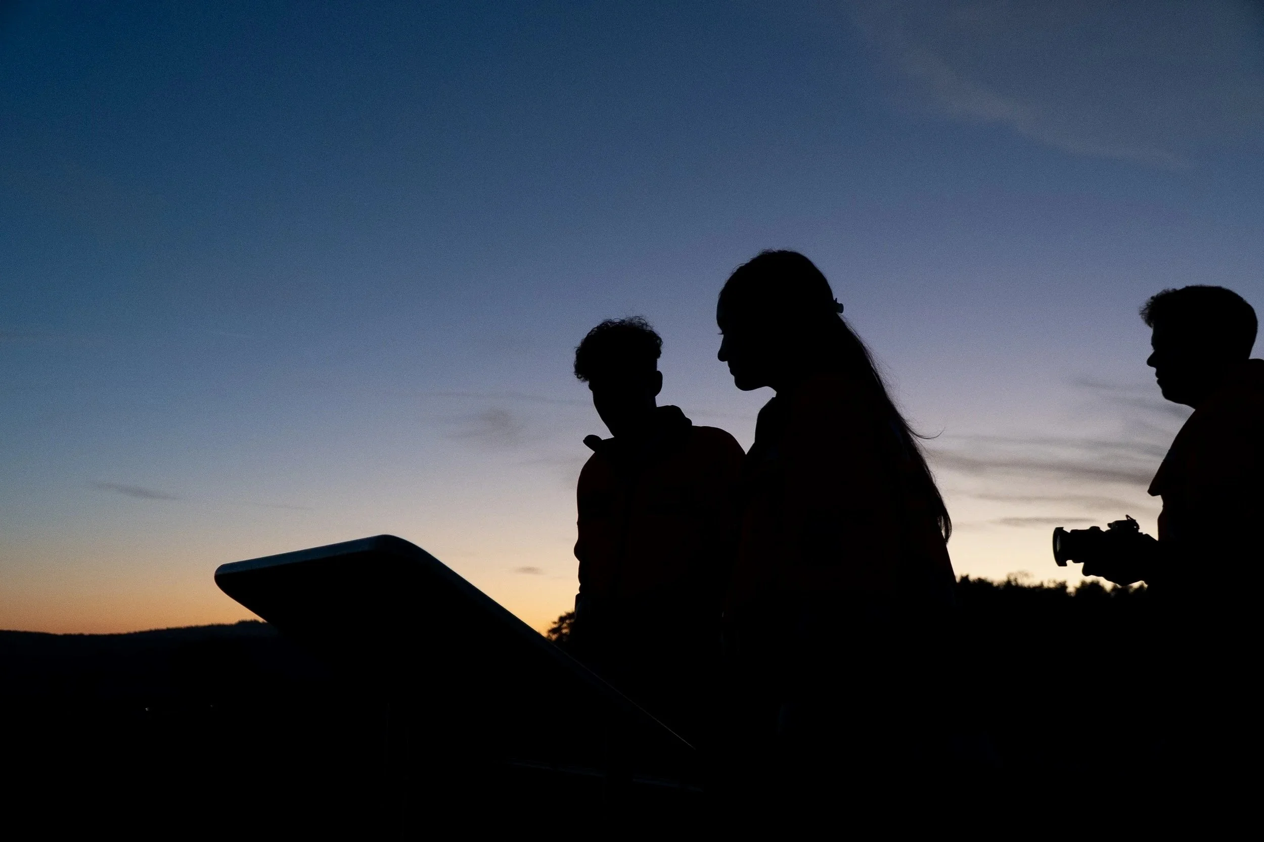 Silhouetten von vier Menschen, zwei Frauen und zwei Männer, stehen bei Sonnenuntergang im Freien und schauen auf mobile Geräte.