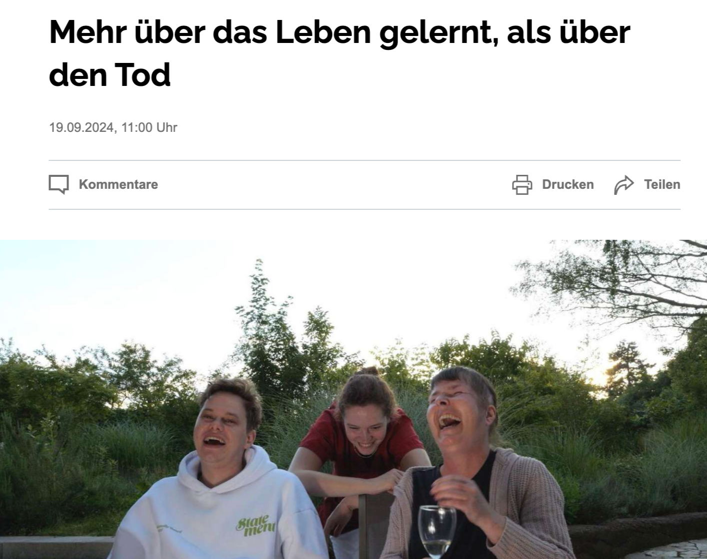 Drei Menschen sitzen im Freien, lachen und scheinen Spaß zu haben, umgeben von Bäumen und Natur.