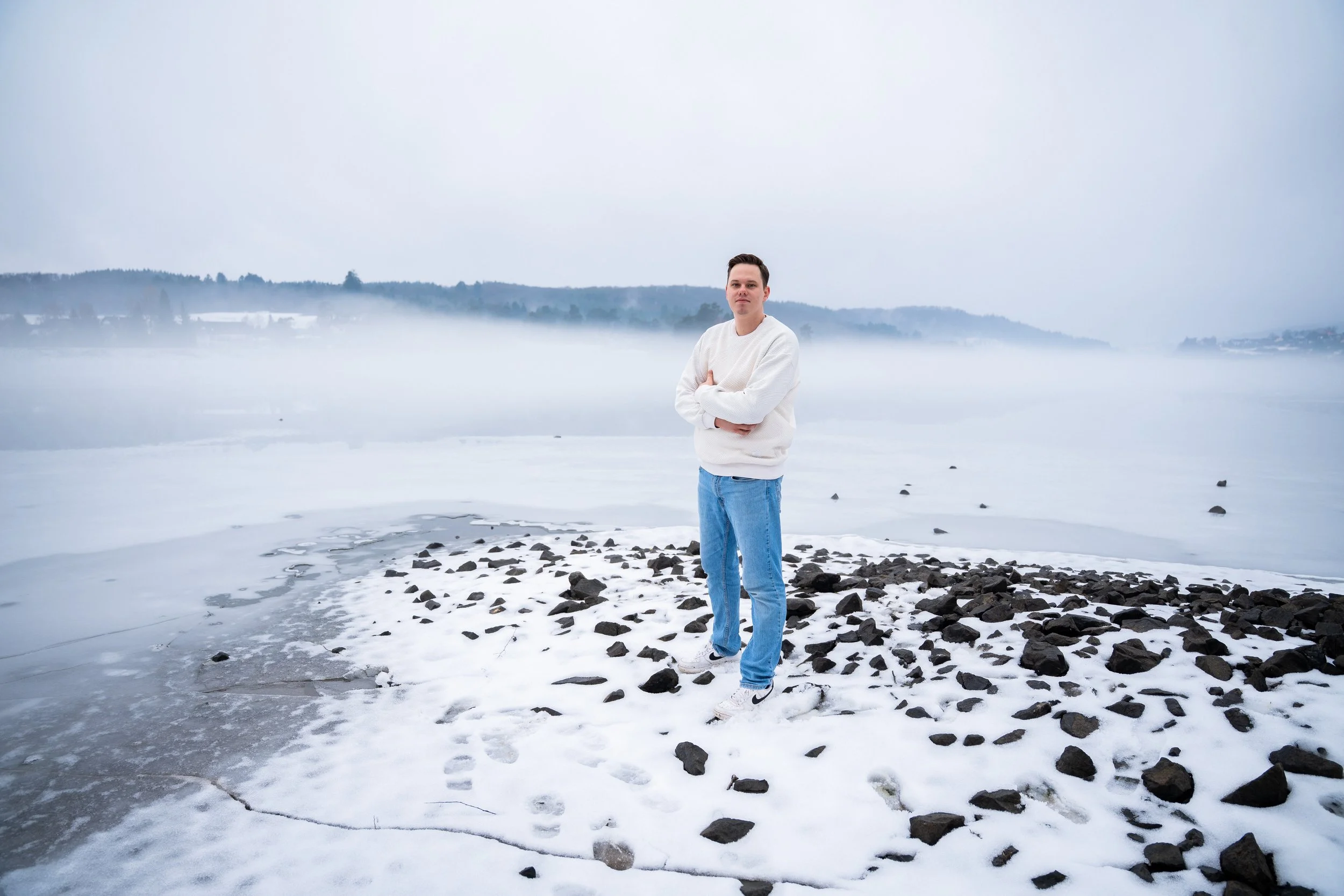 Junger Mann steht an einem eisbedeckten Flussufer, trägt weiße Jacke und blaue Jeans, im Hintergrund Nebel über dem Wasser und bewaldete Hügel.