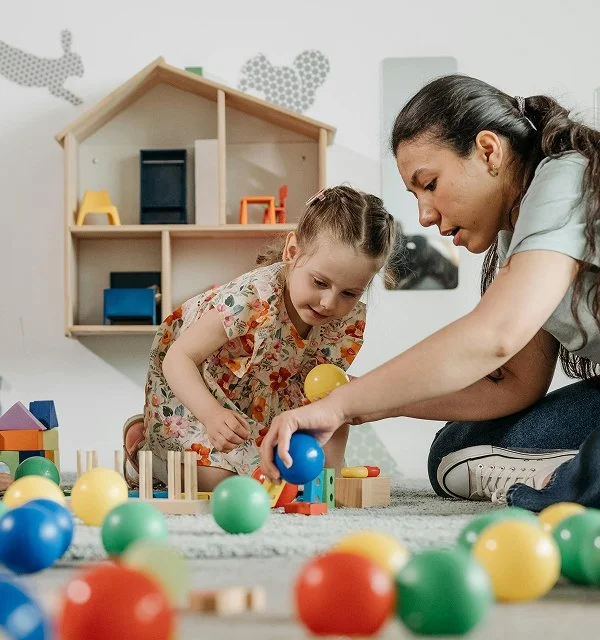 A young girl and a woman playing with colorful balls and wooden toys on the floor in a playroom.