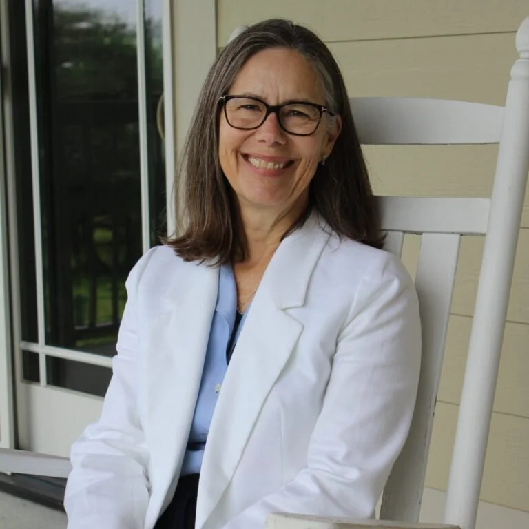 A smiling middle-aged woman with glasses and shoulder-length brown hair, wearing a white coat, sitting on a white rocking chair outdoors near a house with beige siding and a glass door.