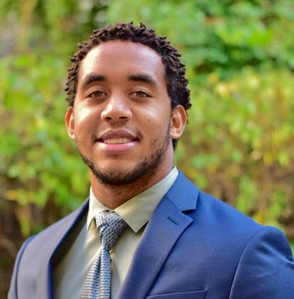 A young man with curly hair and a beard, wearing a blue suit and light-colored dress shirt, standing outdoors with a blurred green background.