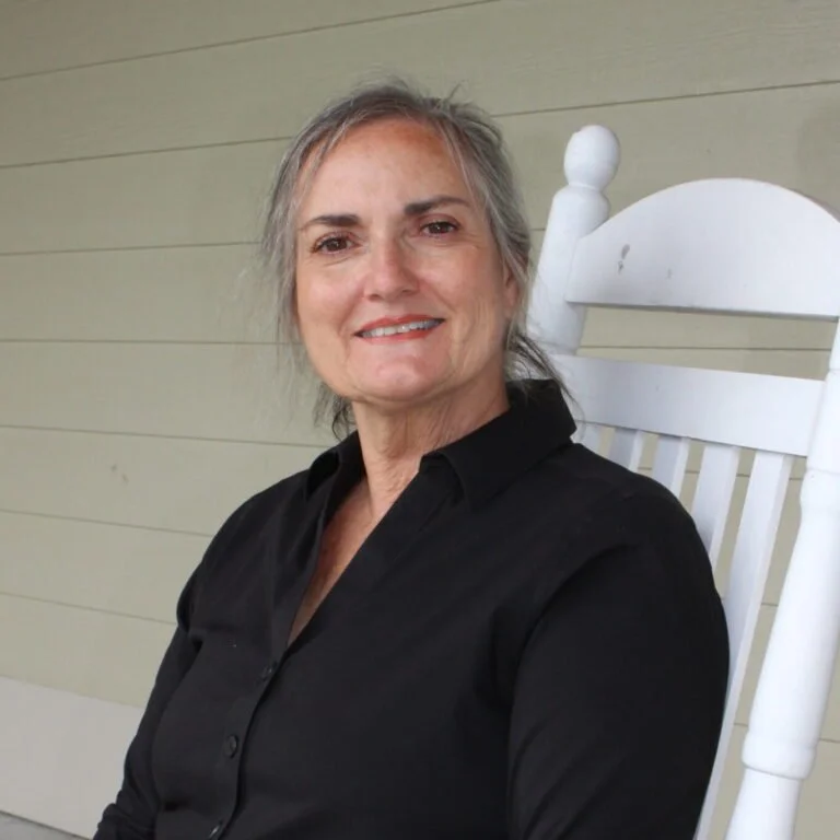 A smiling middle-aged woman with gray hair sitting on a white rocking chair, wearing a black button-up shirt, in front of a light-colored wooden wall.