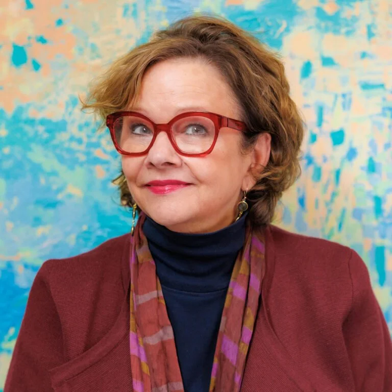 A woman with short, wavy brown hair wearing red glasses, a dark turtleneck, a burgundy blazer, and a multicolored scarf, standing in front of an abstract blue and yellow background.