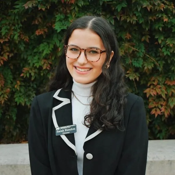 A woman with long dark hair, glasses, and a white turtleneck sweater, wearing a black blazer with white trim, standing outdoors in front of green and brown leafy shrubbery.