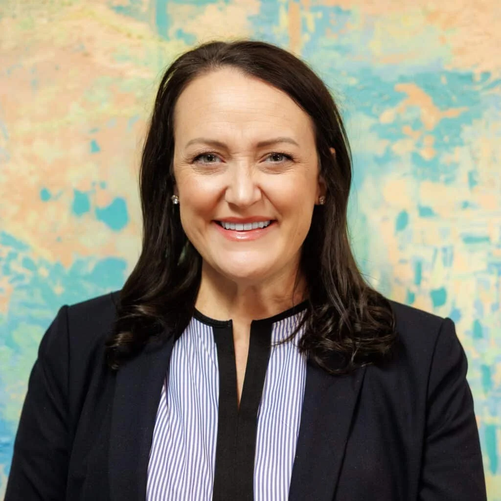A woman with shoulder-length dark hair, smiling, wearing a dark blazer over a blouse with thin vertical stripes and a black collar, standing in front of a colorful abstract background.