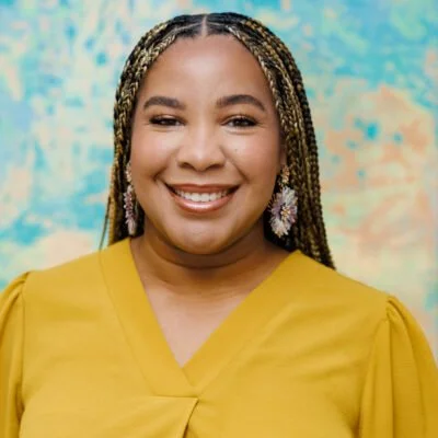 A woman with braided hair smiling, wearing a mustard yellow top and colorful earrings, against a vibrant, abstract background.