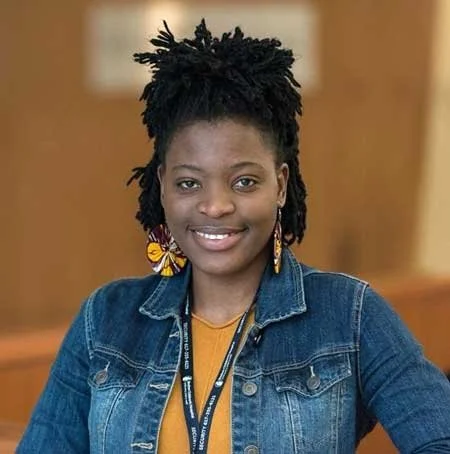 A woman with dark dreadlocks wearing a denim jacket and yellow earrings, smiling at the camera.