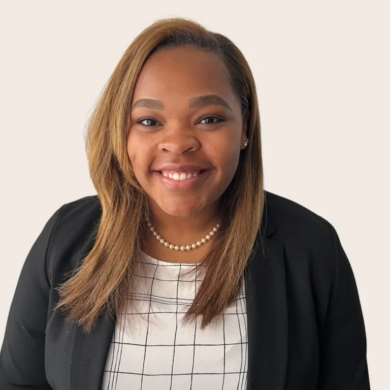 A smiling woman with straight, shoulder-length hair, wearing a black blazer, a white checkered top, and a pearl necklace, standing against a light background.