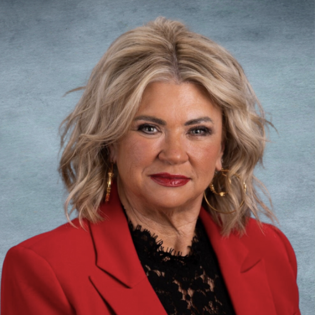 A woman with blonde, wavy hair wearing a red blazer and black lace top, standing in front of a blue background.