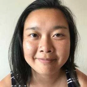 Close-up of a woman with black hair and light skin, smiling softly. She is wearing a striped top and is against a plain background.