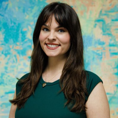 A young woman with long brown hair and bangs, smiling, wearing a dark green top and a necklace, standing in front of a colorful abstract background.