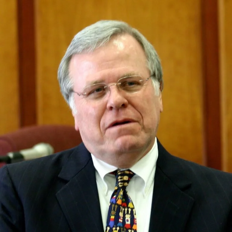 A middle-aged man with gray hair, glasses, wearing a black suit, white shirt, and a colorful patterned tie, sitting in a room with wooden paneling