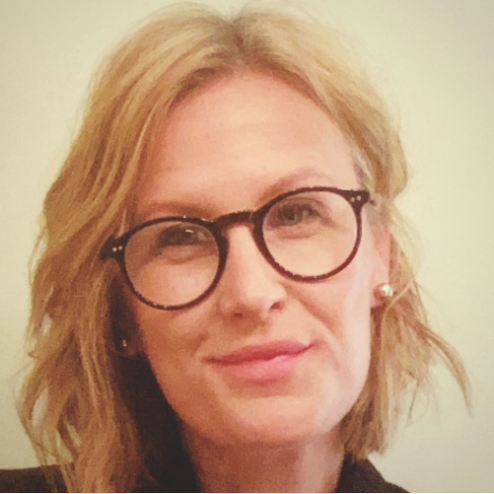 A woman with blonde hair, wearing glasses with round frames, and pearl earrings. She is smiling slightly and facing the camera.