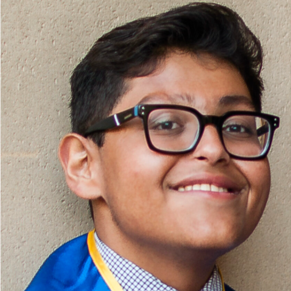 Close-up of a young person with black hair, glasses, and a blue shirt smiling against a plain wall.