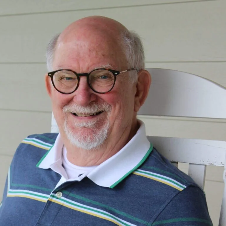 A smiling elderly man with glasses, white hair, and a beard sitting on a white wooden chair, wearing a striped polo shirt.
