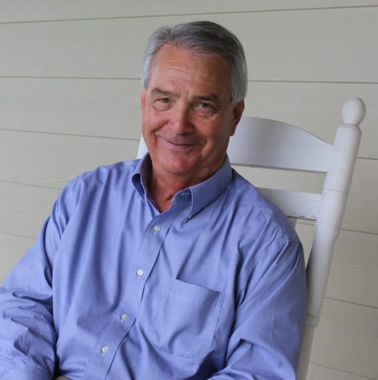Older man with gray hair smiling, sitting on a white wooden rocking chair, wearing a light blue button-down shirt, on a porch with horizontal beige siding.