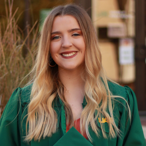 A young woman with long blonde hair, wearing a green graduation gown and holding a diploma, smiling outdoors.