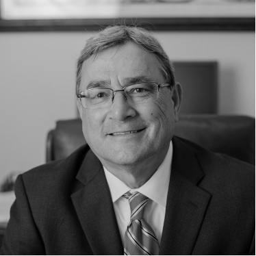 A grayscale photo of a smiling middle-aged man in a suit and tie, wearing glasses, sitting in an office chair.