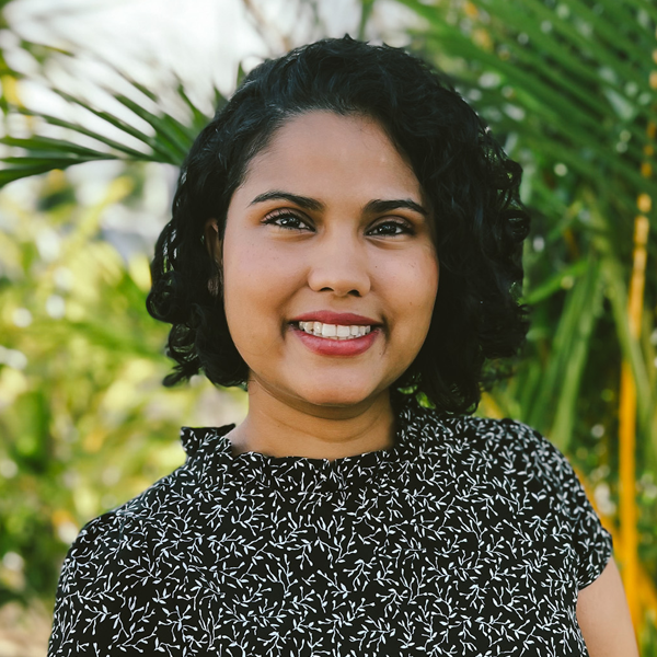A woman with short, curly black hair smiling outdoors in front of green plants, wearing a black patterned top.