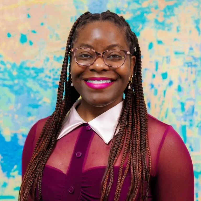 A woman with long braided hair wearing glasses, bright pink lipstick, a white collared shirt, and a maroon top, smiling in front of an abstract blue and yellow background.
